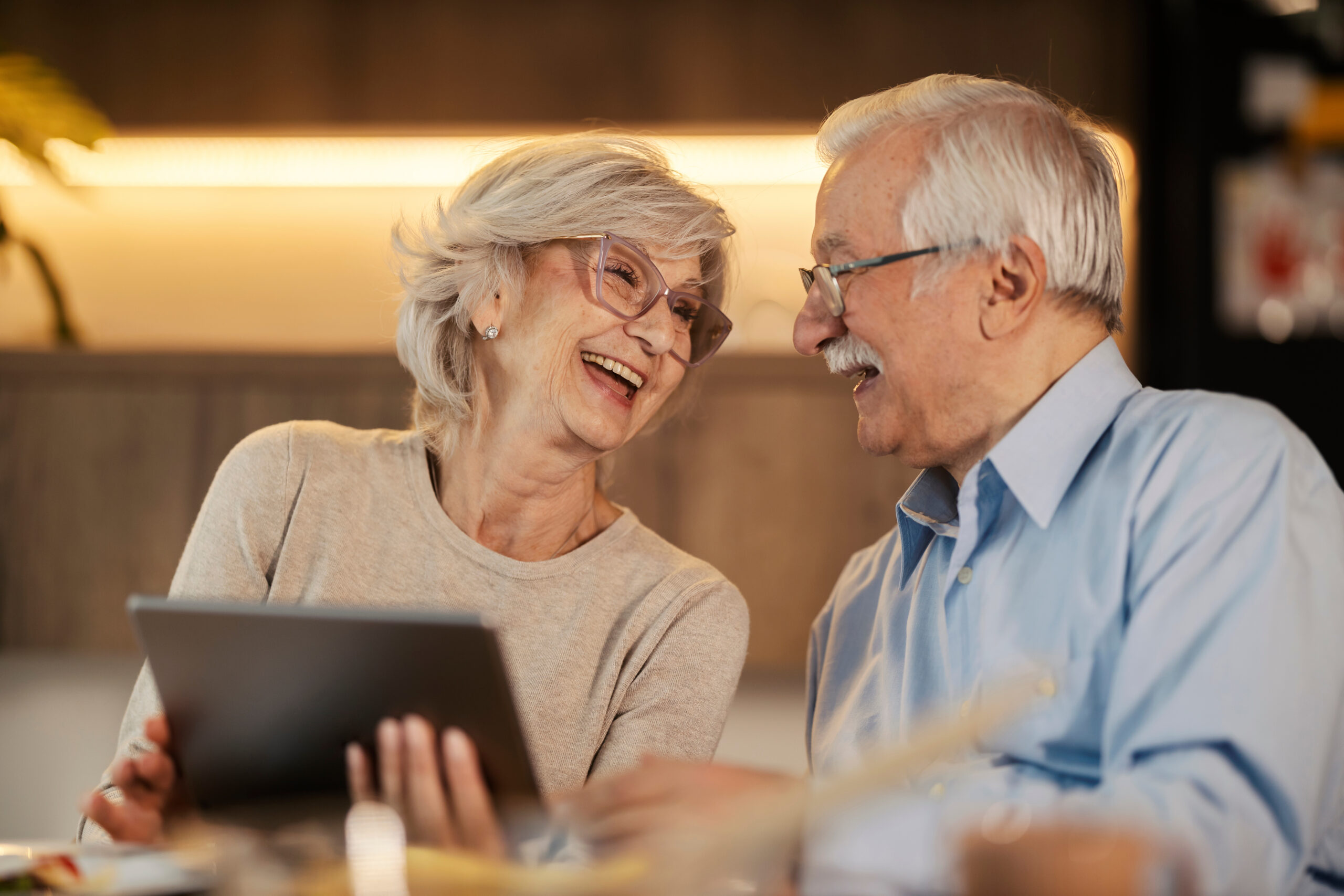 Cheerful-senior-couple-laughing-at-dining-table-and-holding-tablet