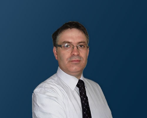 A person wearing a white dress shirt and a black tie with small white dots stands against a solid dark blue background.