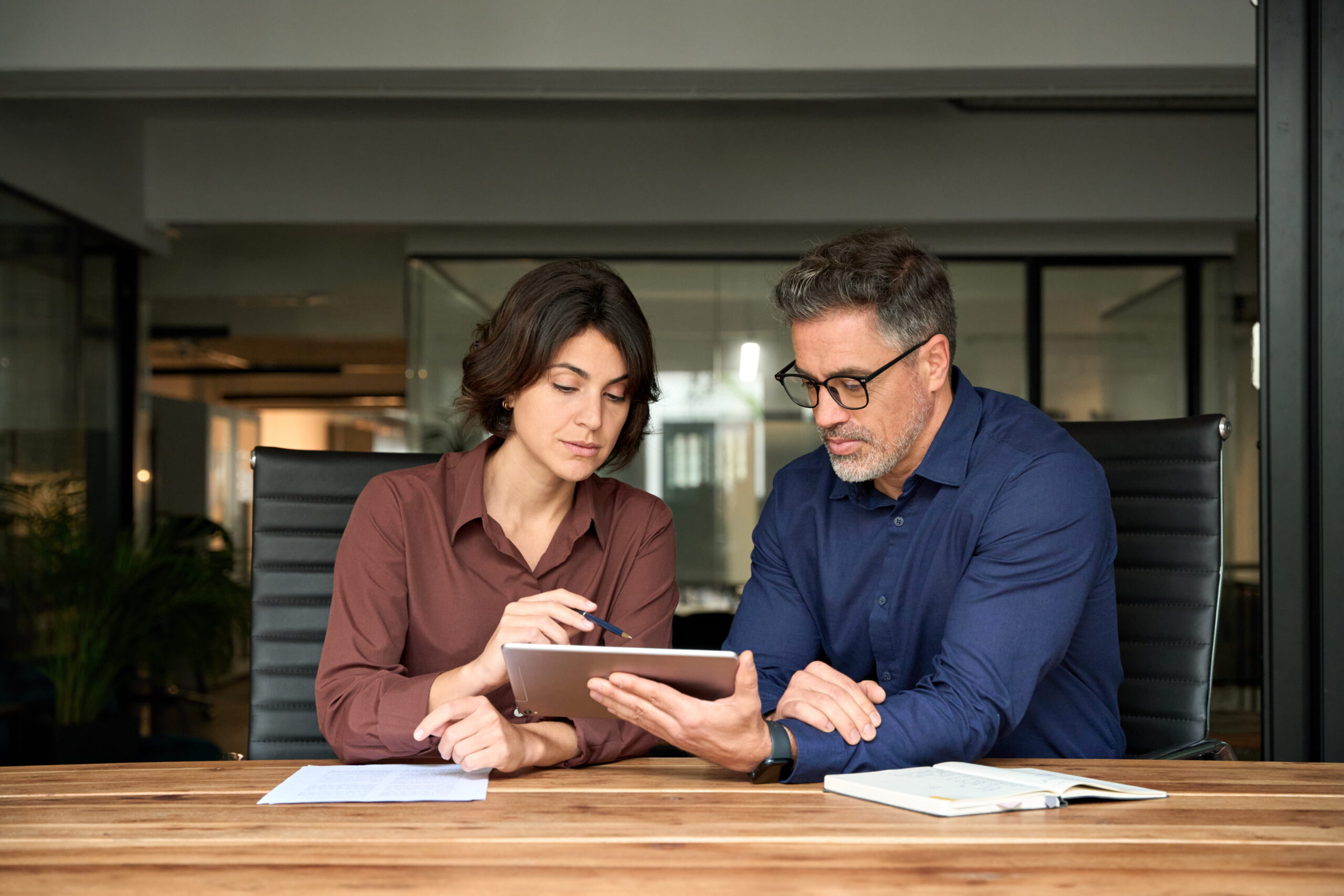 Two people seated at a wooden table in a modern office setting, looking at a tablet together
