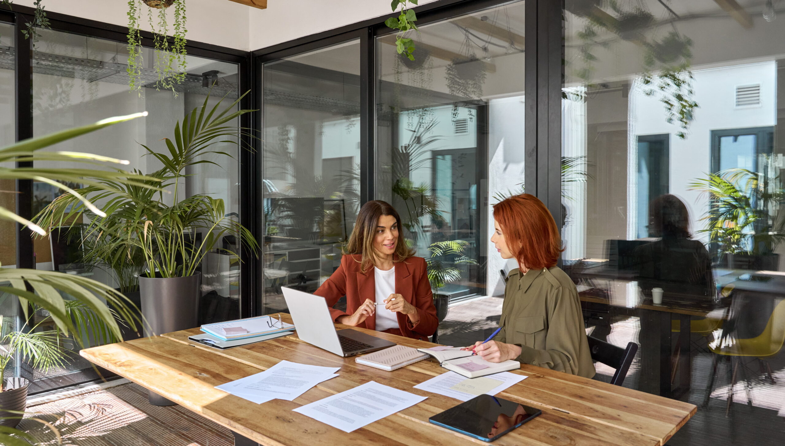 Two people are seated at a wooden table in a bright, plant-filled office environment