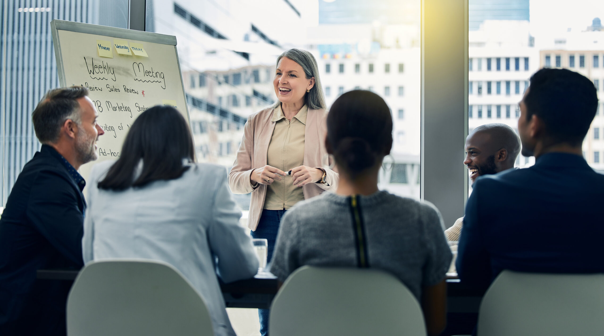 A woman standing at the front of a meeting room near a large window, speaking to a group seated around a table.