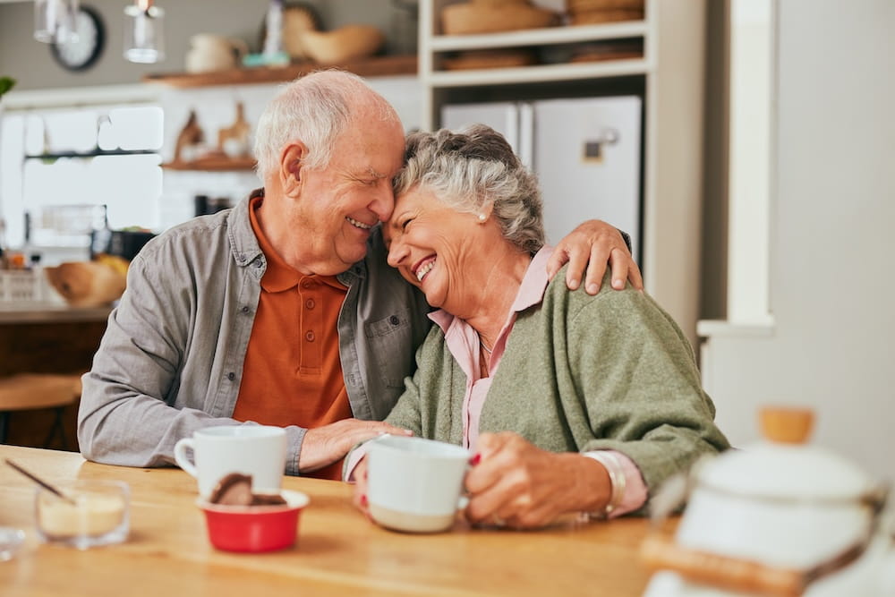 A happy older couple embrace sat at a table with cups of tea on the table in front of them