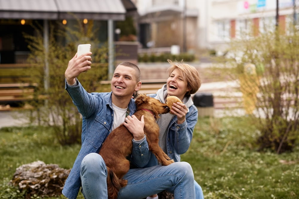 A happy couple take a selfie sat on a lawn holding a puppy