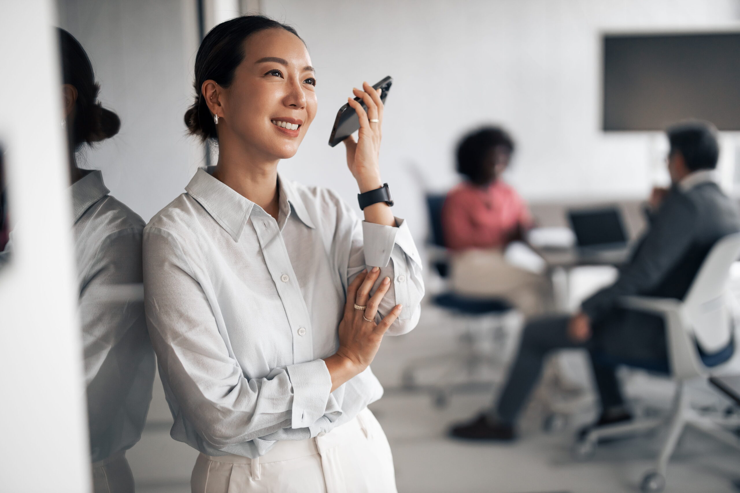 A woman smiles while on a phone call. Two people sit in the background having a discussion.