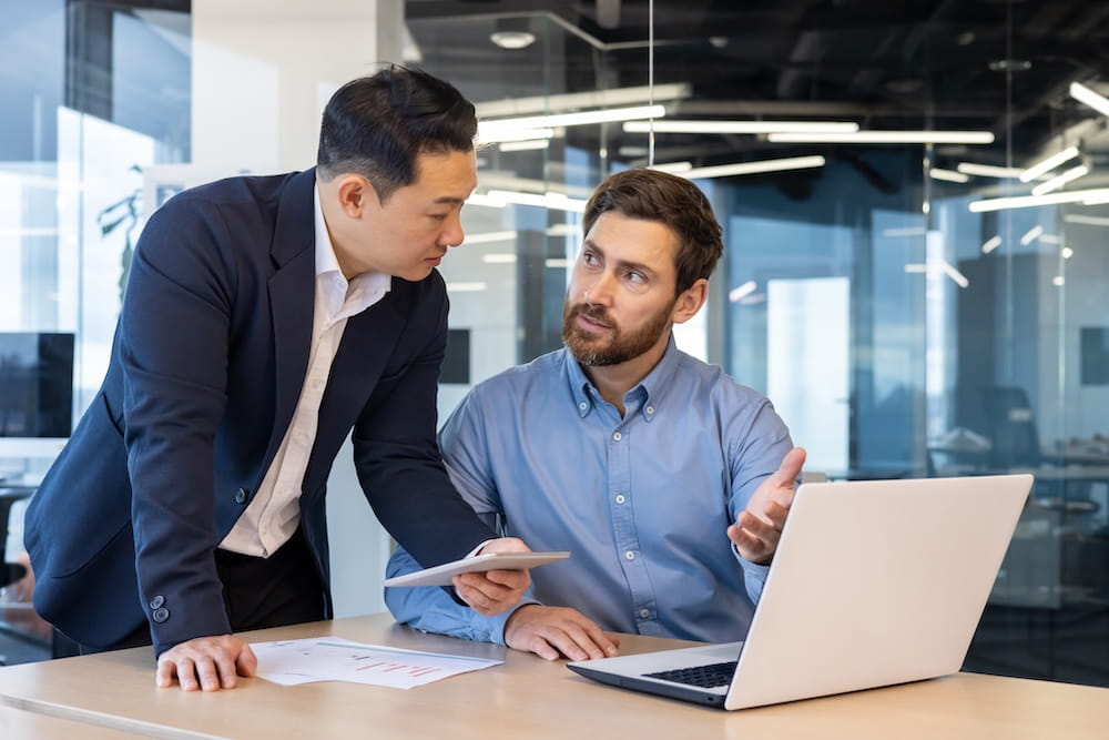 Two men at a desk in an office, one is sat and is presenting some work on a laptop