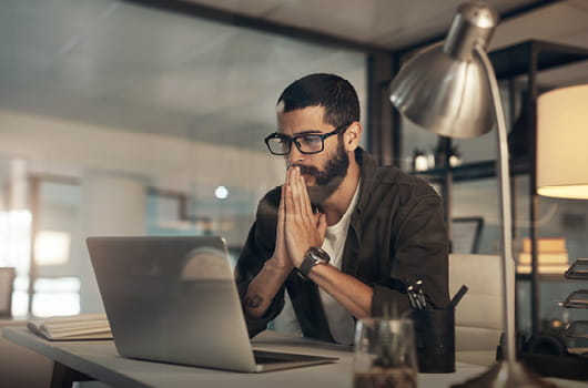 A man seated at a desk in an office, hands clasped in front of his face while looking at an open laptop, with a desk lamp and notebooks nearby.