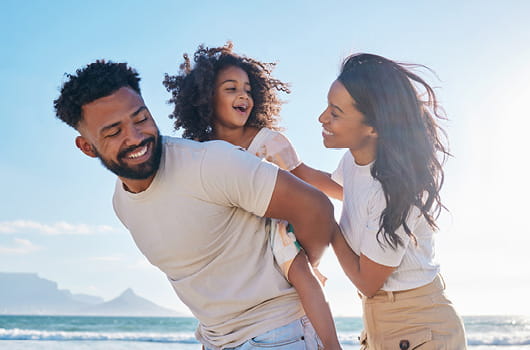 Three people outdoors near the sea, with man carrying a little girl on his back while a woman walks alongside.
