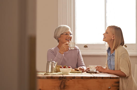 Two women sitting at a wooden kitchen table near a window, holding cups while plates of food and a teapot are placed on the table.