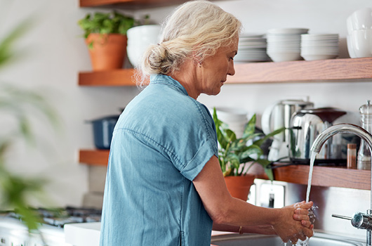 A person in a blue short-sleeved shirt washes hands under a kitchen tap, with wooden shelves, white bowls and potted plants in the background.