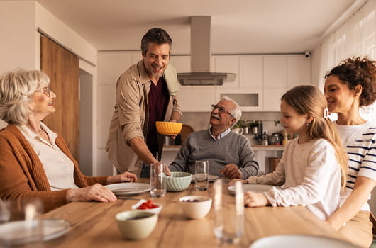 Five people gathered around a wooden dining table in a kitchen, with one person handing a bowl to another while others sit nearby with plates, glasses, and food on the table.