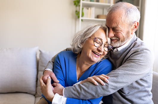 An elderly couple sitting closely on a sofa, with one person wrapping their arms around the other, in a living room setting with cushions and shelving in the background.