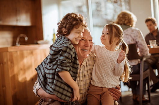 An elderly gentleman embracing two children in a home setting, with wooden kitchen units and a dining table with seated people visible in the background.