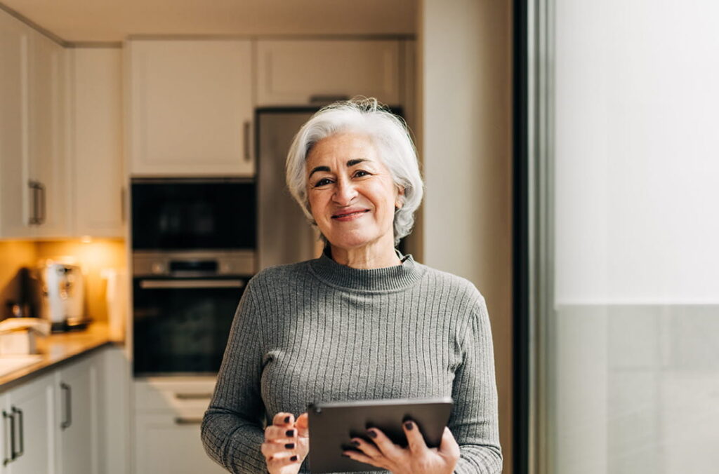 A woman standing in a modern kitchen holding a tablet, with built-in appliances, countertops, and a large window visible in the background.