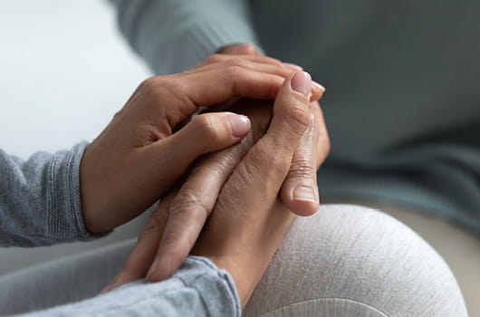 Close-up view of two pairs of hands clasped together, with one person holding the other's hands while seated indoors.